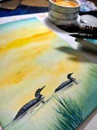 watercolor painting of 2 loons on water golden light shown on artist's desk
