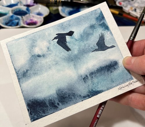 hand holding watercolor painting of ravens in flight against a misty background