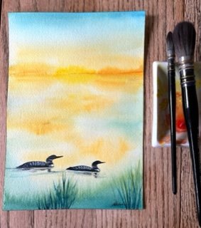 watercolor painting of 2 loons on water golden light shown with paintbrushes